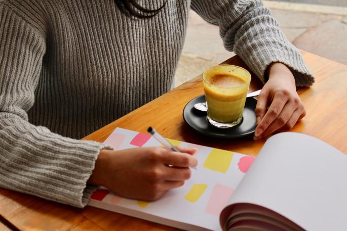 Person sitzt mit Getränk am Tisch und schreibt in Notizbuch